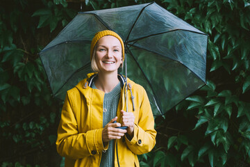 Happy woman wearing yellow raincoat holding transparent umbrella green leaves wall. Thoughtful...