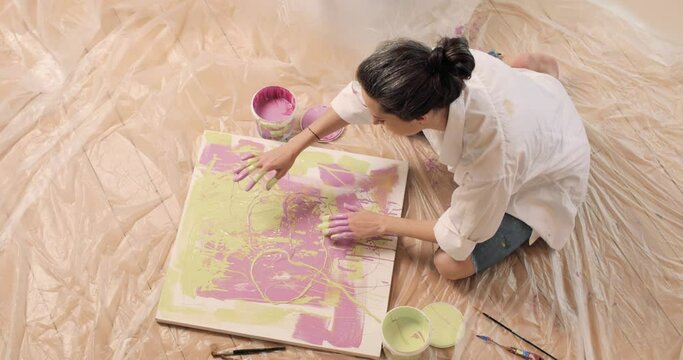 Top view of woman artist in the white shirt smearing paint with her hands on the canvas on the floor in the studio. Art creativity concept.