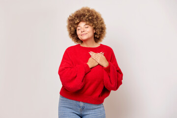 Cute young woman with afro style hair wearing red sweater stands on light background, she is happy...