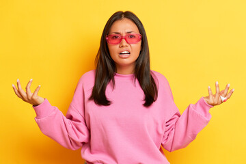 Nice brunette woman in pink sunglasses and sweatshirt standing with hands up on yellow studio backdrop, she is dissatisfied with something, copy space, high quality photo