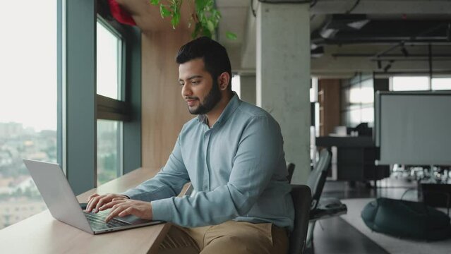 Pensive Indian man working on laptop near window in the office