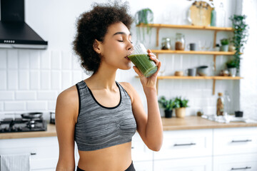 Healthy eating, veganism, raw food. Lovely happy healthy african american or hispanic woman, stand in the kitchen, drinking healthy fortified organic smoothie from glass cup, taking care of health