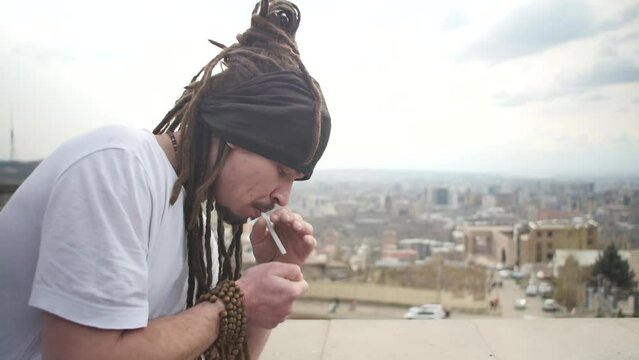 A guy with dreadlocks, beads and bracelets on his arm sets fire to a cigarette in his mouth, inhaling cigarette smoke into his lungs. A young man in a white T-shirt and dreadlocks in a bun smokes