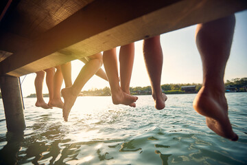 Rear view of legs of group of friends by lake. Four young friends sitting on jetty with their legs hanging down to the water on a summer day. Holiday, togetherness, lifestyle concept.