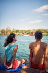 Fototapeta premium Man and woman sitting by water on wooden jetty. Rear view of couple. Woman stretching. Togetherness, holiday, lifestyle concept.