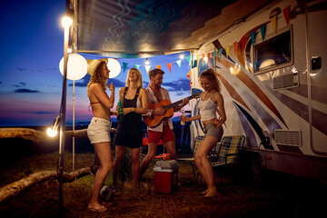 Friends camping at summertime with trailer in nature. Young man playing guitrar, women dancing and talking. Togetherness, holiday, weekend, fun, lifestyle concept.