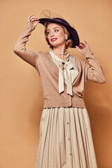 Attractive woman wearing old-style clothes smiling, touching hat and looking away over beige background
