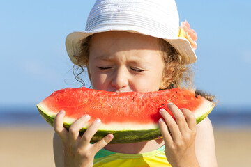 cute little girl eats ripe juicy red watermelon on the beach, coast, seashore. the concept of summer kid holidays, seasonal fruits and berries. closeup portrait