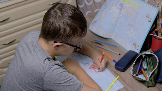 Sad Boy in glasses Drawing Country of Russia on World Map with a Red Pencil. Schoolboy is sitting at the table in bedroom, doing homework in geography. War in Ukraine. Concept of the Russian invasion.
