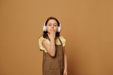 Obraz premium Caucasian adorable little child girl dressed in stylish khaki dress, listening to music on wireless headphones, blowing out and sending air kisses looking at camera, isolated over beige background.