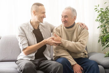 Happy two generations male family old senior mature father and smiling young adult grown son enjoying talking chatting bonding relaxing having friendly positive conversation sit on sofa at home.