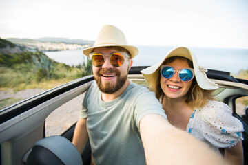 Happy beautiful couple in love taking a selfie portrait driving a convertible car on the road at vacation. Rental cars and vacation