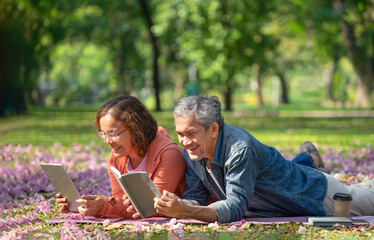 asian senior married couple enjoy outdoors leisure time in the park,they are lying on grasses,a man reading a book and woman playing digital tablet,concept elderly people lifestyle,resting,relaxing