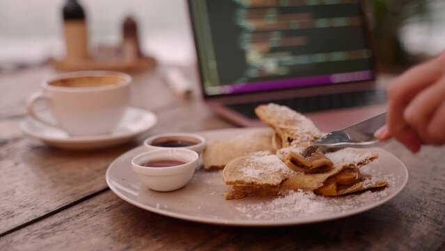 Young woman developer has lunch break eating food while working on laptop in outdoor cafe. Female freelancer having pankakes and coffee at coffeeshop for breakfast. IT girl eats her food at cafe