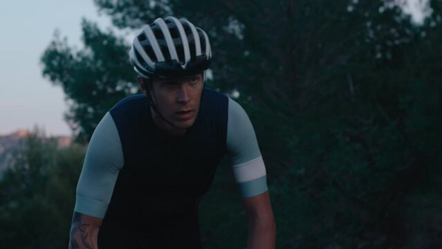 Fit and concentrated male cyclist on heavy cardio training. Young professional athlete climb up mountain hill on carbon road bike. Cinematic cycling during evening training session