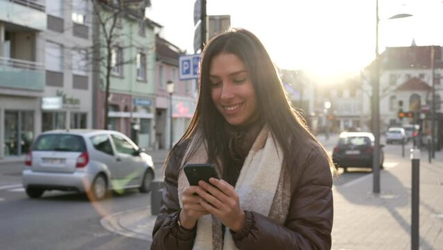Joyful young woman holding smartphone in city street smiling. Adult female 20s girl liking content online looking at phone in urban town sidewalk