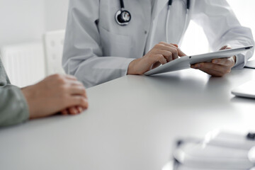 Doctor and patient sitting at the desk in clinic office. The focus is on female physician's hands using tablet computer, close up. Medicine concept