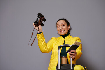 Delightful woman traveler adventurer, going for trip, posing with vintage binoculars and boarding pass, smiling cutely looking at camera, standing near her yellow suitcase on isolated gray background