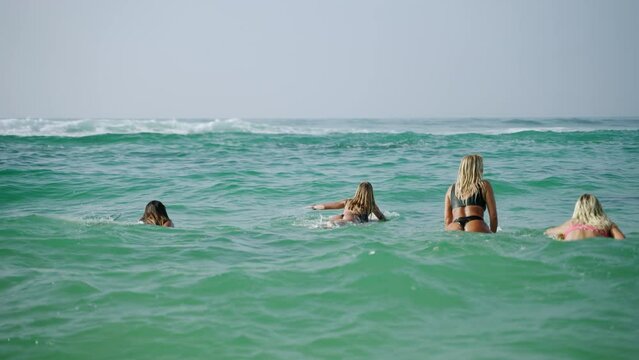 Young multiracial women surfers paddling surfboards and riding waves in the sea at tropical beach at sunrise. Girl friends enjoy outdoor lifestyle and water sport surfing on summer vacation back view