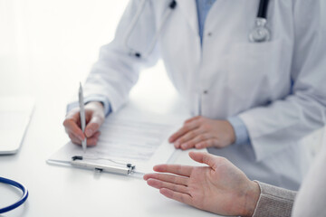 Doctor and patient sitting at the table in clinic. The focus is on female physician's hands filling up the medication history record form or checklist, close up. Medicine concept
