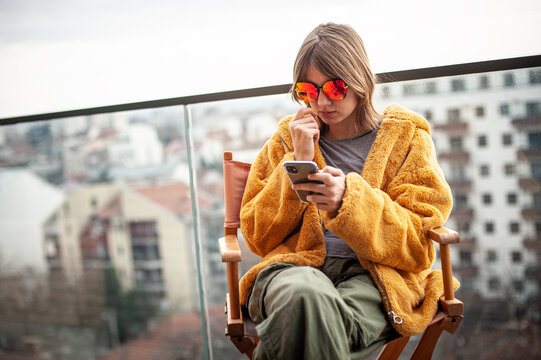 Beautiful young girl in a yellow fur coat and red glasses is sitting in a director's chair on the terrace of a modern building. Use smartphone, social media influence, taking picture, 