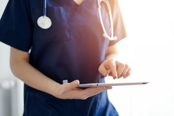 Doctor woman using tablet computer while standing near panorama window in clinic, close up. Physician or surgeon at work. Medicine concept