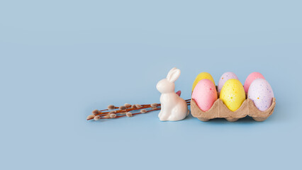 Happy easter decoration. White bunny decorative statuette, willow branch, basket with colorful eggs. Minimal festive composition on pastel light blue background. Copy space, place for text, front view