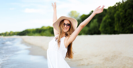Fototapeta na wymiar Happy smiling woman in free happiness bliss on ocean beach standing with a hat, sunglasses, and rasing hands. Portrait of a multicultural female model in white summer dress enjoying nature