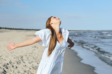 Happy smiling beautiful woman is on the ocean beach in a white summer dress, open arms