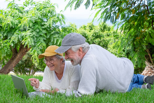 Happy senior couple lying in the meadow in the park making video call using laptop computer together. Video call concept.