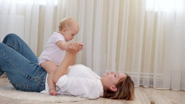Happy loving millennial caucasian mom lie on back on floor in light cozy bedroom lift up toss in air small baby daughter or son.Caring mum laugh cuddle kiss cute infant babe have fun enjoy motherhood.