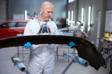 Middle-aged man in protective gloves prepares a car body part for painting