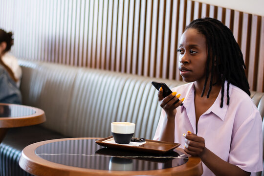 Upset african-american woman speaks a voice message on her phone while sitting in a modern cafe and drinking coffee. Use of gadgets, smartphones, artificial intelligence, voice assistant