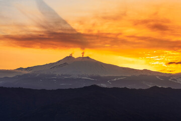 mysterious landscape of great erupting volcano with smoke from craters and snow on slopes in orange light of sunset. eruption of vulcan
