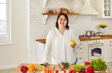 Overweight young woman cooking healthy food in modern kitchen. Cheerful plus size woman standing at table preparing salad of fresh vegetables at home. Healthy eating, organic vegetarian food