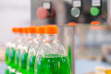 Row of pet bottles with green lemonade and orange caps on conveyor belt of automatic liquid filling machine at plastic exhibition - close up. Manufacturing, industry and technology equipment concept
