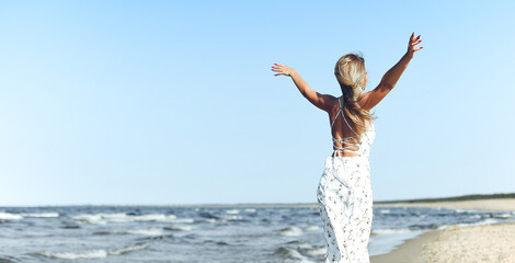 Fototapeta na wymiar Happy blonde beautiful woman on the ocean beach standing in a white summer dress, open arms
