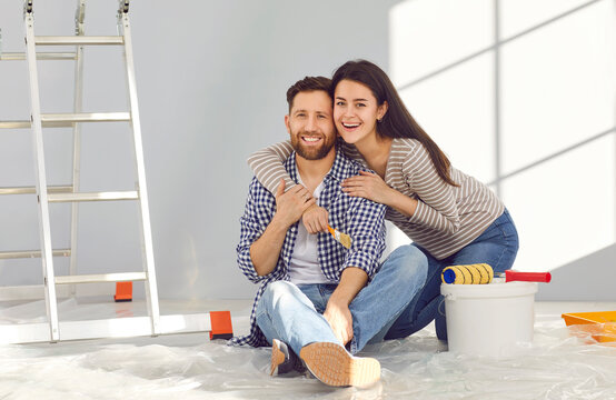 Cheerful married couple sitting on floor in their new house. Happy wife hugging her husband and smiling at camera in freshly painted room. Couple painting walls with paint rollers and brush at home