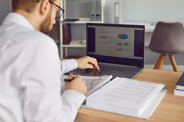 Male accountant sitting at office desk, working with business documents, checking financial data, using laptop computer, and looking at budget infographic pie and bar charts on computer screen