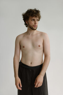 A topless curvy guy in trousers with pierced nipples looks thoughtful
