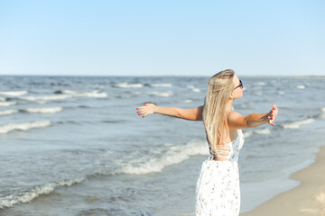 Happy blonde beautiful woman on the ocean beach standing in a white summer dress and sun glasses, open arms