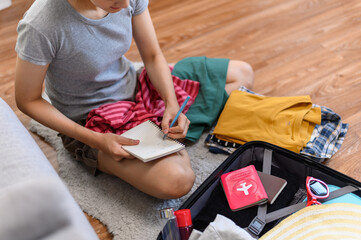 Young beautiful Asian woman using checklist prepare arrange clothes and equipment before go travel oversea after covid pandemic. New normal travel lifestyle concept.