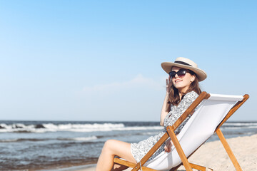 Happy brunette woman wearing sunglasses and hat relaxing on a wooden deck chair at the ocean beach