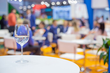 Close up view: glass of water, champagne or wine on white table against blurred business meeting, conference, seminar background. Corporate and communication concept