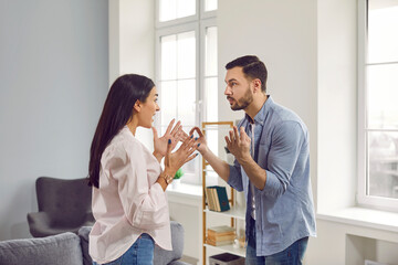 Angry young couple standing at home and arguing in the living room shouting on each other. Frustrated unhappy husband and wife quarreling. Bad marriage,relationships, quarrel and divorce concept. - obrazy, fototapety, plakaty