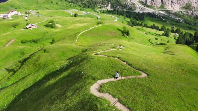 Alone fashionable dressed woman merry walking by mountain path on Green hills
over Passo Gardena in Dolomite Alps, Italy. 4K Aerial flying drone shot.