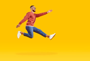 Naklejka na ściany i meble Excited positive man runs and jumps with fast speed trying to achieve his goals. Full length happy man in casual clothes levitating in air taking step on orange background. Side view in profile.