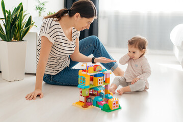 Laughing mother and little daughter playing colorful blocks, constructing tower, sitting on warm...