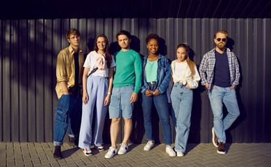 Fototapeta premium Young people in casual everyday clothes leaning on street wall. Group portrait of six happy relaxed beautiful multiethnic fashion models posing in shirts, tees, jumpers, jeans and denim jackets
