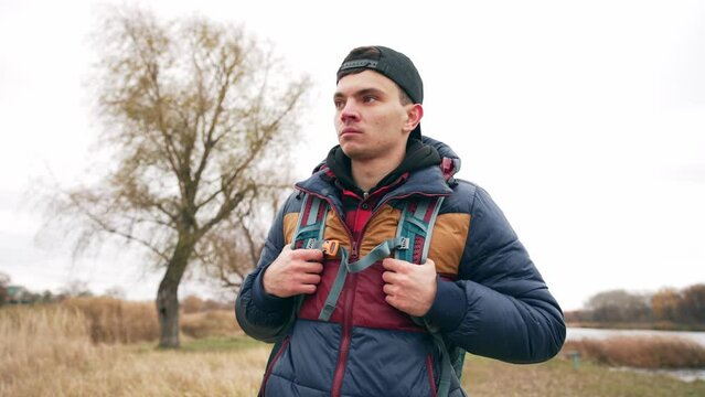 Portrait of young guy tourist backpacker in cap with smartphone finds route on map hiking in autumn. He looks around in search of right path. Hike, tourism, travel, explore wild nature concept.
